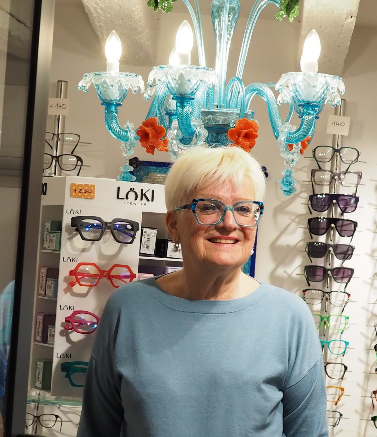 Signora con occhiali turchesi sotto il lampadario in vetro soffiato di Ottica Silvana, tra montature ed espositore LÖKI
