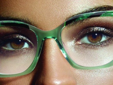 Primo piano di occhi castani con trucco bronzo dietro una montatura in acetato verde trasparente con venature scure