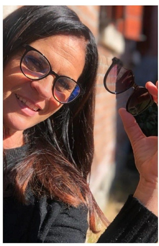 Donna sorridente con occhiali tondi neri mostra una clip da sole scura in piena luce davanti a un muro di mattoni